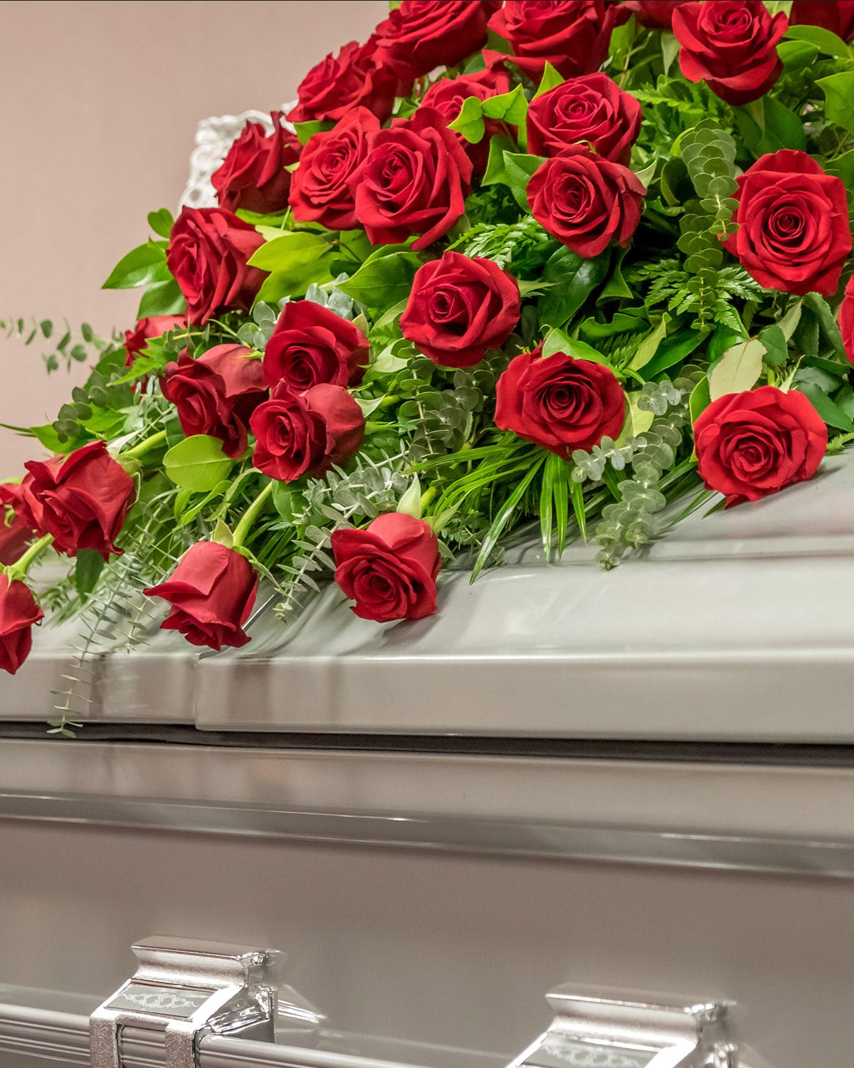 A large bouquet of red roses and green foliage rests on a closed, silver-gray casket adorned with shiny metallic handles, set against a soft pink background.