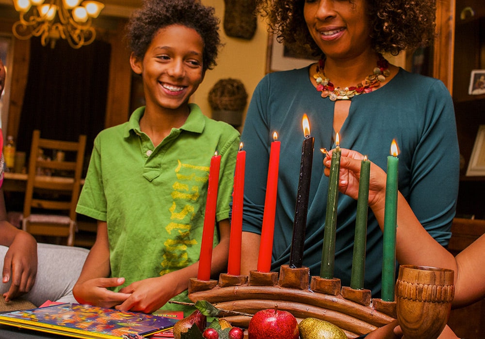 A smiling boy and a woman light Kwanzaa candles in a ceremonial holder, surrounded by festive decorations and warm indoor lighting.