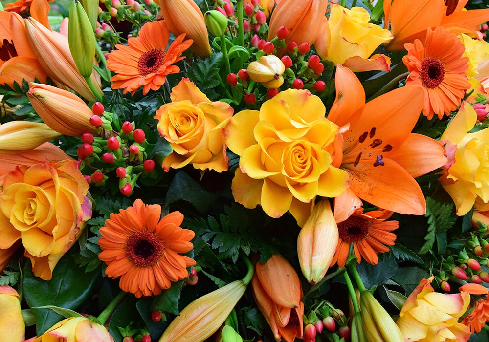 A vibrant floral arrangement featuring blooming yellow roses, orange lilies, and gerbera daisies, accented by unopened buds and green foliage, creating a vivid and lush display.