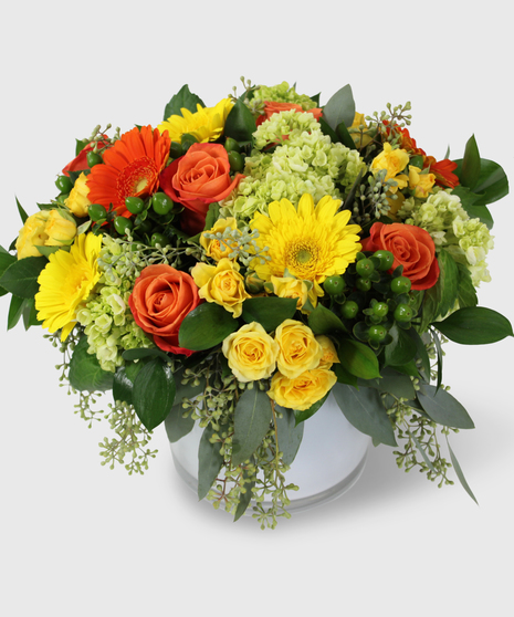 Bright yellow, orange, and green flowers with greenery in a white cylinder vase.