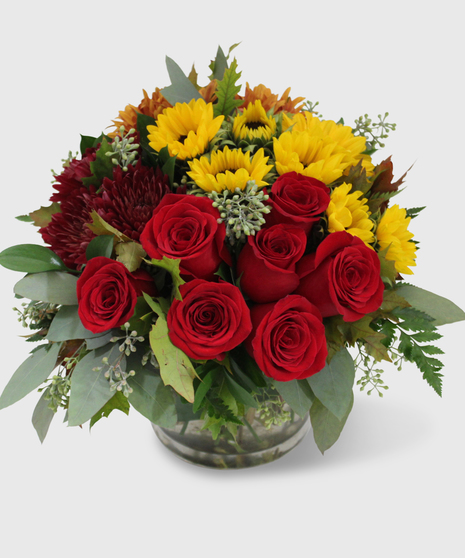 A compact vase with red roses, red cremons, bronze mums, mini sunflowers and lush greenery