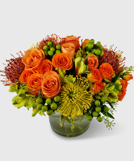 A unique fall flower arrangement designed in a short compact cylinder vase with orange and yellow pincushion protea, orange roses, orange spray roses, green berries and more