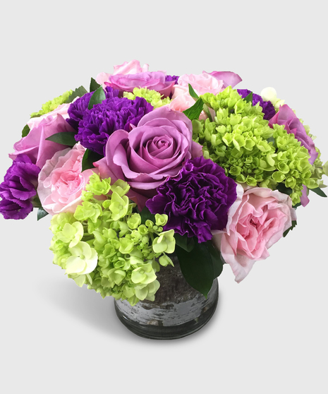 Purple roses, carnations, pink roses, green hydrangea and more in a cylinder vase.