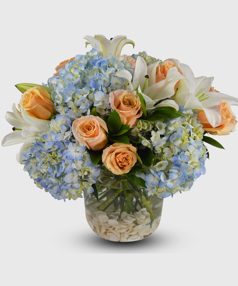 A summer vase with sea shells on the bottom designed with blue hydrangea, peach roses and white lilies