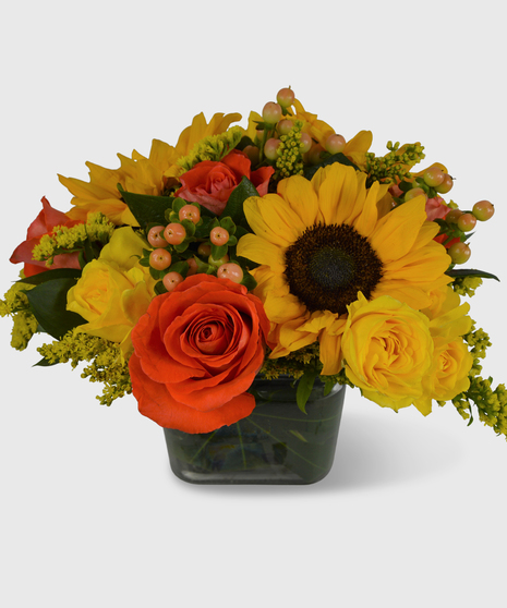 A fall bouquet designed in leaf-lined block vase with sunflowers, orange roses, yellow spray roses and orange berries