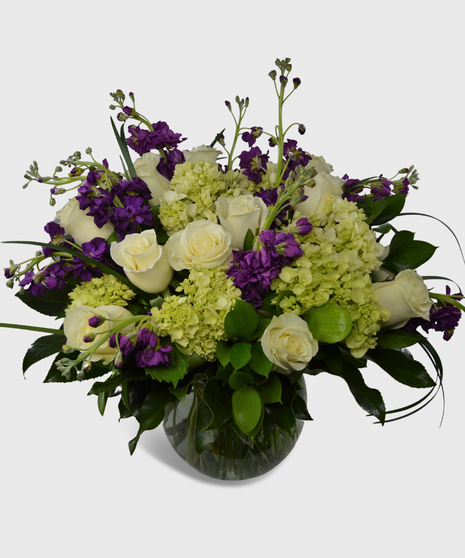 Purple stock, creme roses, green hydrangea and lush greenery designed in a clear glass vase