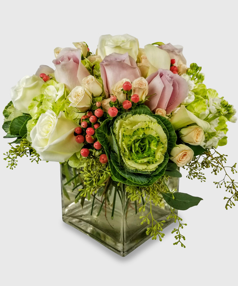 A unique fall bouquet with cream and blush roses, green kale, orange berries and seeded eucalyptus in a heavy block vase