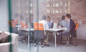 People sitting around a conference table, working on laptops in a modern office with glass walls and an abstract wall design, surrounded by high-rise buildings outside.