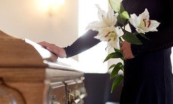 A person in black attire holding white lilies rests a hand on a wooden casket in a softly lit room, suggesting a funeral or memorial service.