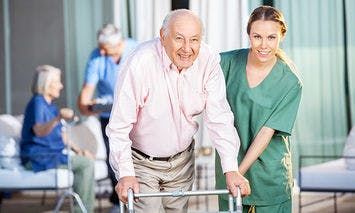 An elderly man uses a walker, aided by a smiling caregiver in green scrubs, in an outdoor setting with other elderly individuals sitting and standing in the background.